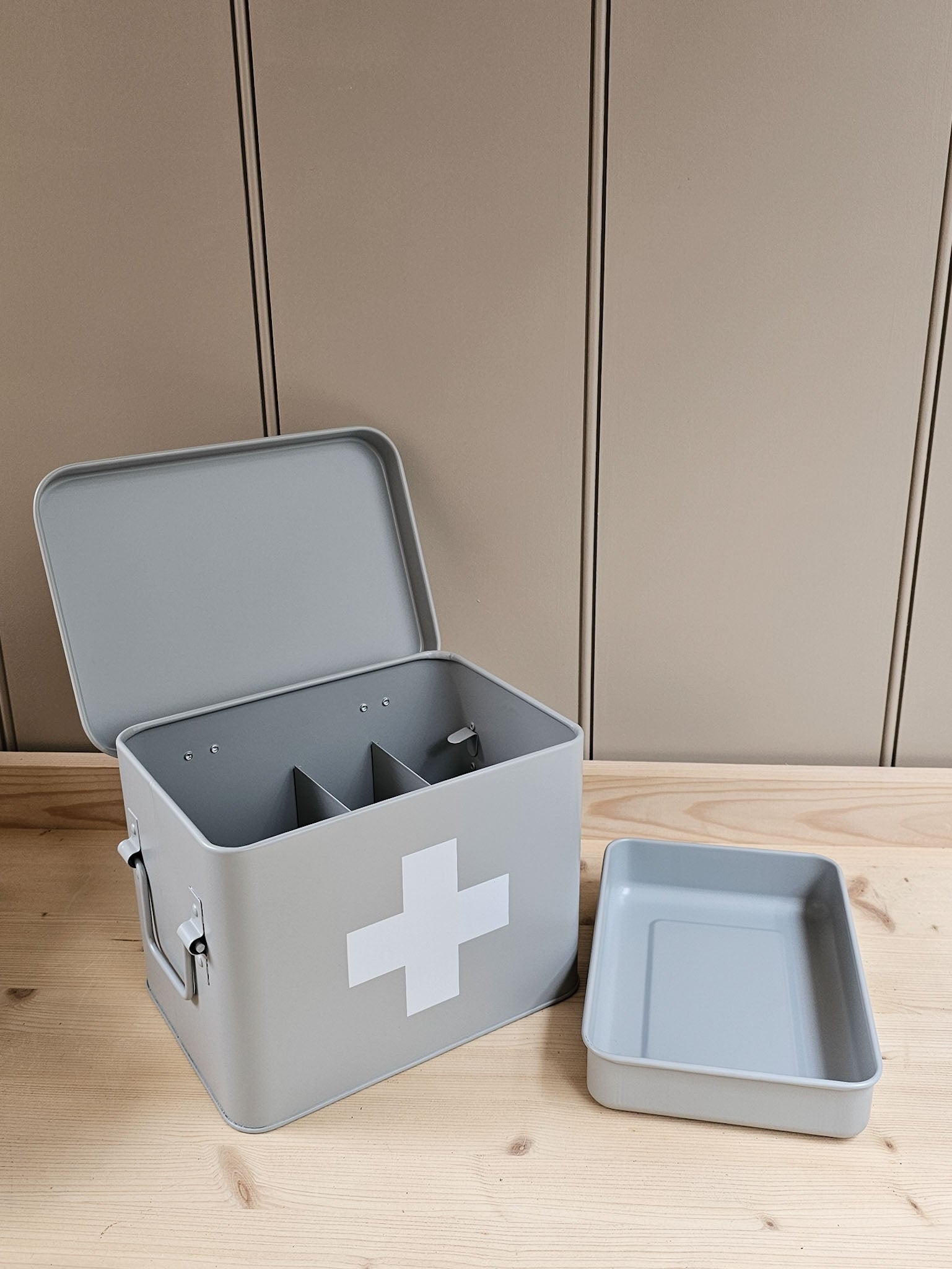 A light gray metal first aid box with an open lid, featuring a white cross symbol on the front and two interior compartments. The matching lid is placed beside it on a light wood surface against a beige paneled background.