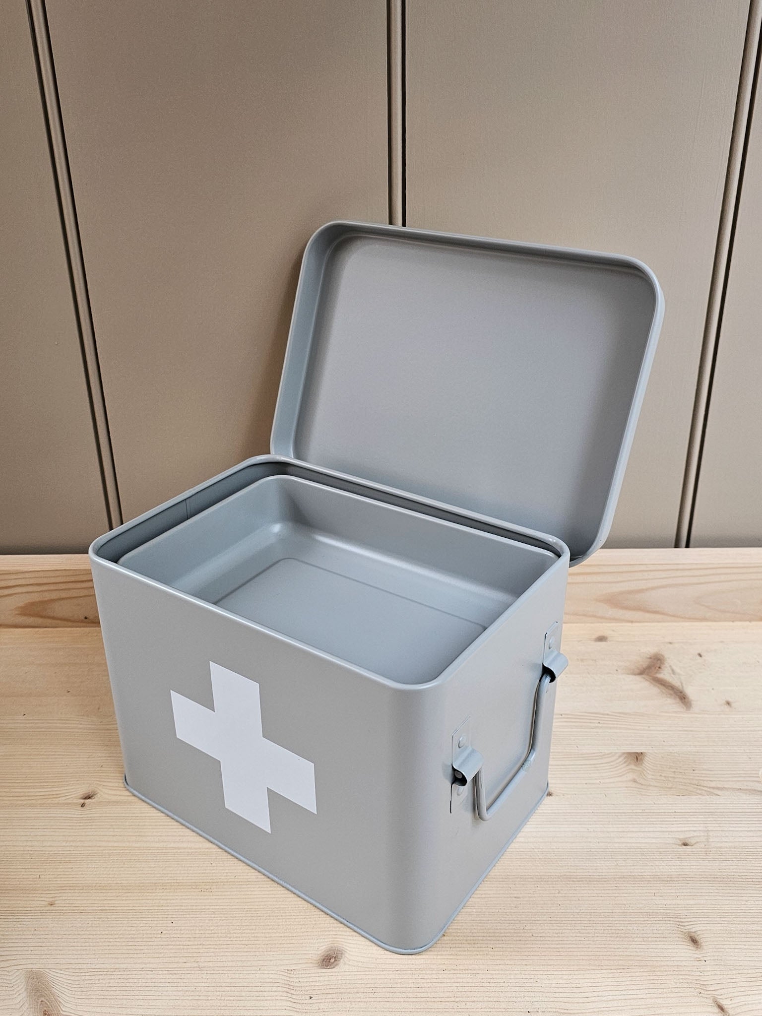 A gray metal first aid box with its lid open, featuring a white medical cross on the front, set on a light wood surface against a beige paneled background.