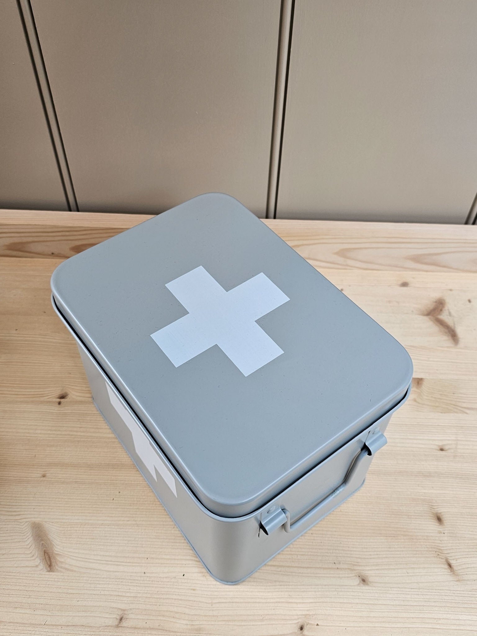 A gray metal first aid box with a white cross symbol on the lid, resting on a light wood tabletop against a beige paneled background.