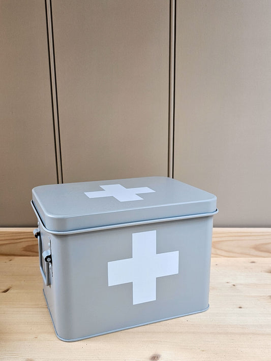 A rectangular light gray metal first aid box with a white cross symbol on the lid and front, sitting on a light wood surface against a beige paneled background.