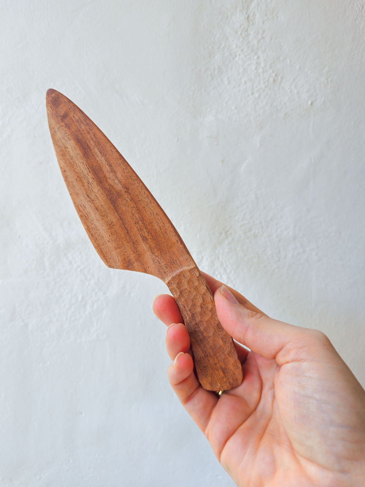 A hand holding a small wooden knife with a smooth, polished blade and a textured, hammered handle against a light, textured off-white wall.
