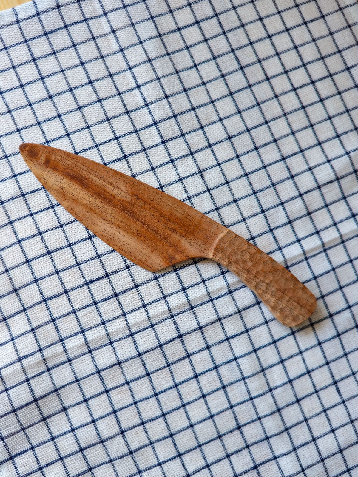 A handcrafted wooden knife with a textured, hammered handle and a smooth blade, resting diagonally on a white kitchen towel with a blue grid pattern.