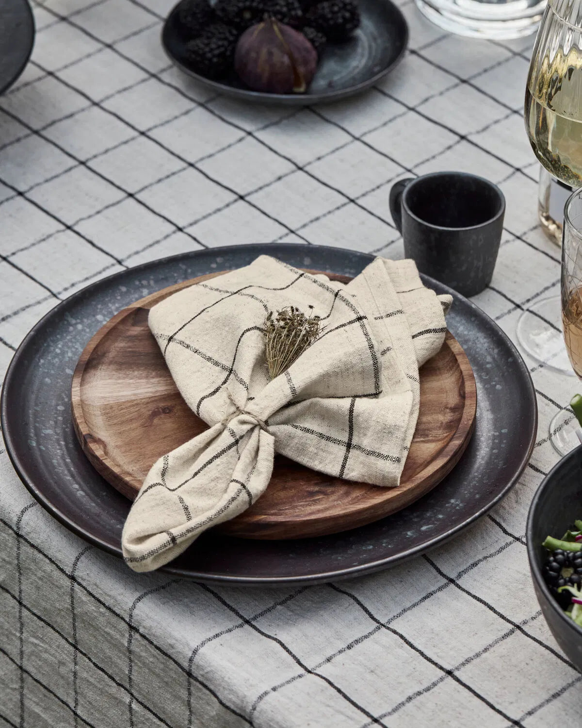 A white and black checked napkin is laying on a wooden plate placed on a dark grey ceramic plate, within a table setting. The table is covered with a tablecloth with the same design as the napkin. 