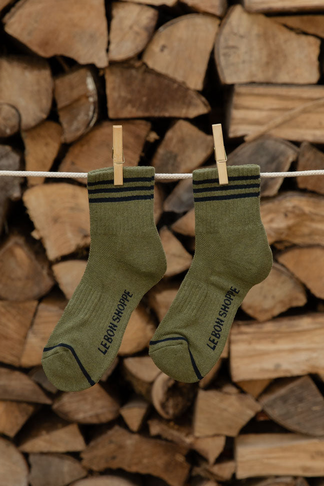 Le Bon Shoppe Girlfriend Socks in Sage with black stripes hanging on a line against stacked logs