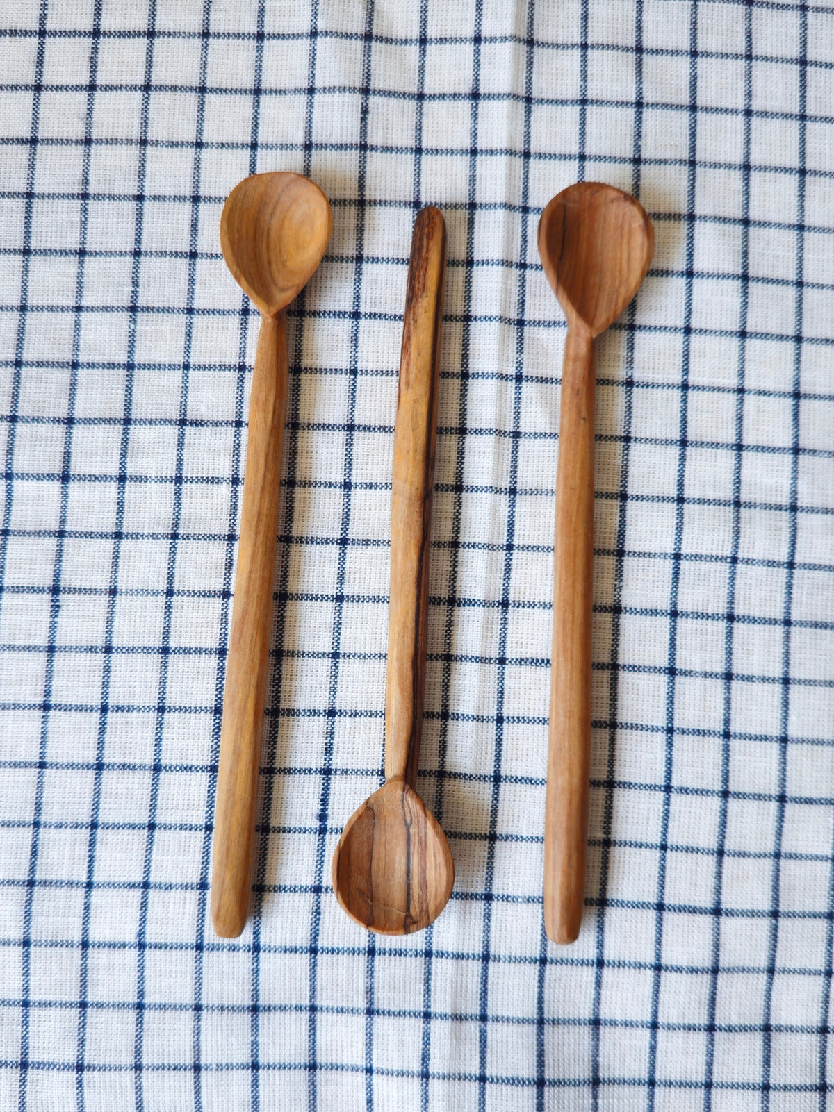 Three handcrafted wooden spoons of varying sizes and types (spoon, teaspoon, soup spoon) arranged vertically on a white and navy blue checkered kitchen towel.