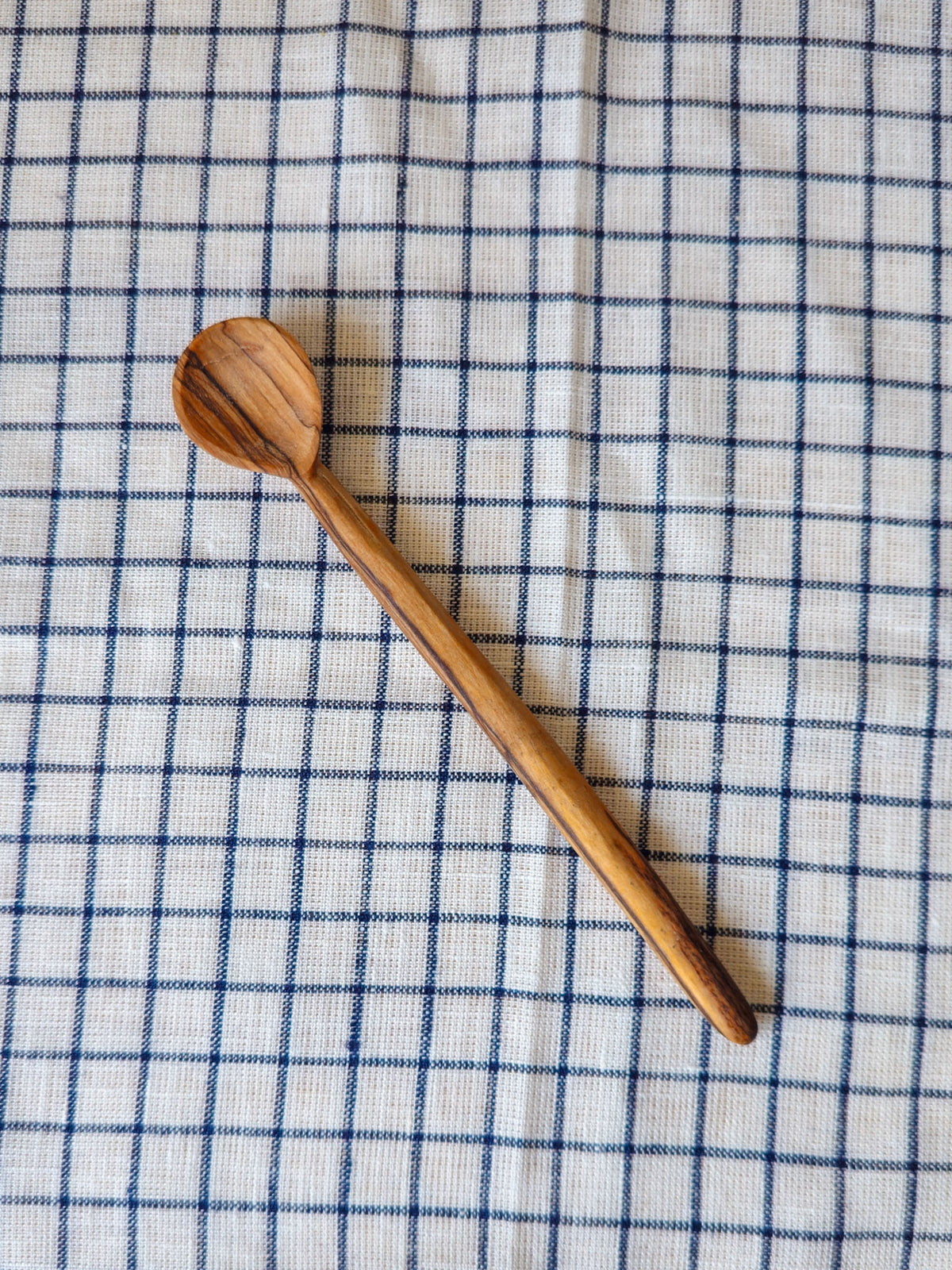 A single wooden spoon with a natural striped wood grain pattern, placed diagonally on a white linen cloth with a blue grid pattern.