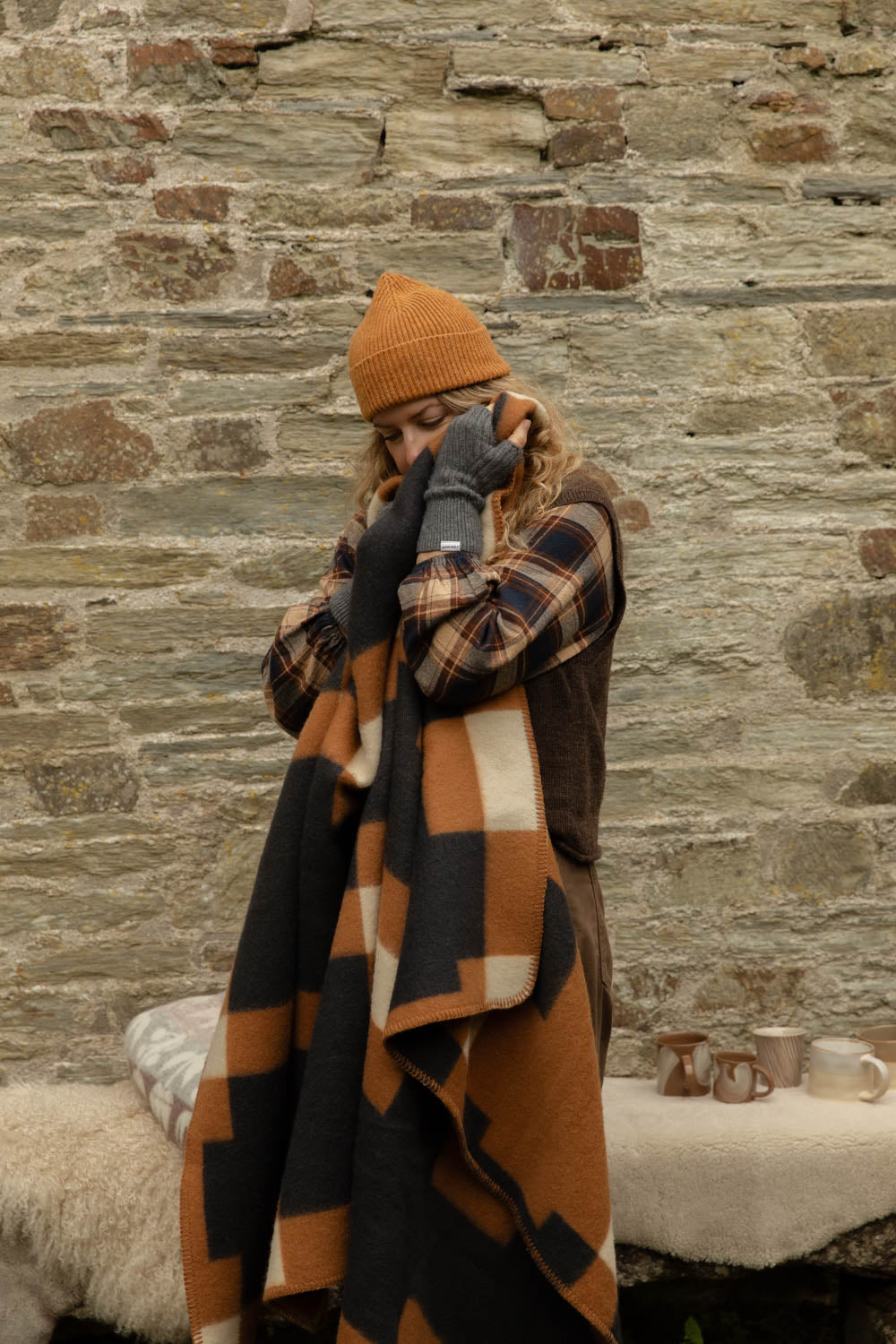 Woman in a mustard ribbed beanie wrapped in an orange, black and cream block-check wool blanket, standing by a stone wall