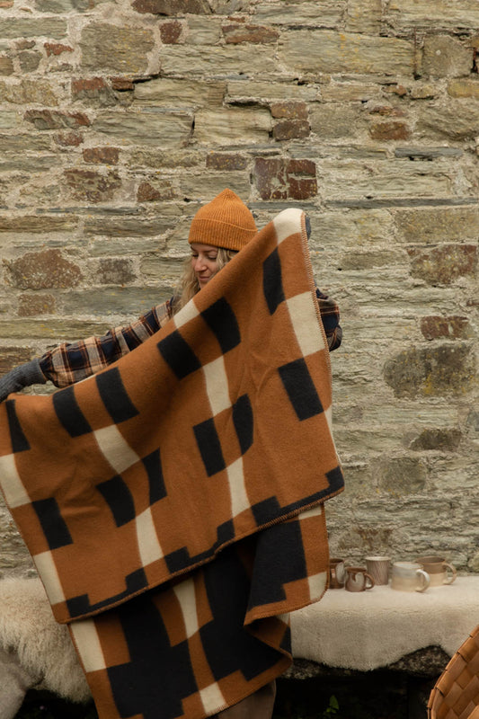 Woman in a mustard ribbed beanie holding open a lambswool blanket in a bold orange, black and cream block check against a stone wall