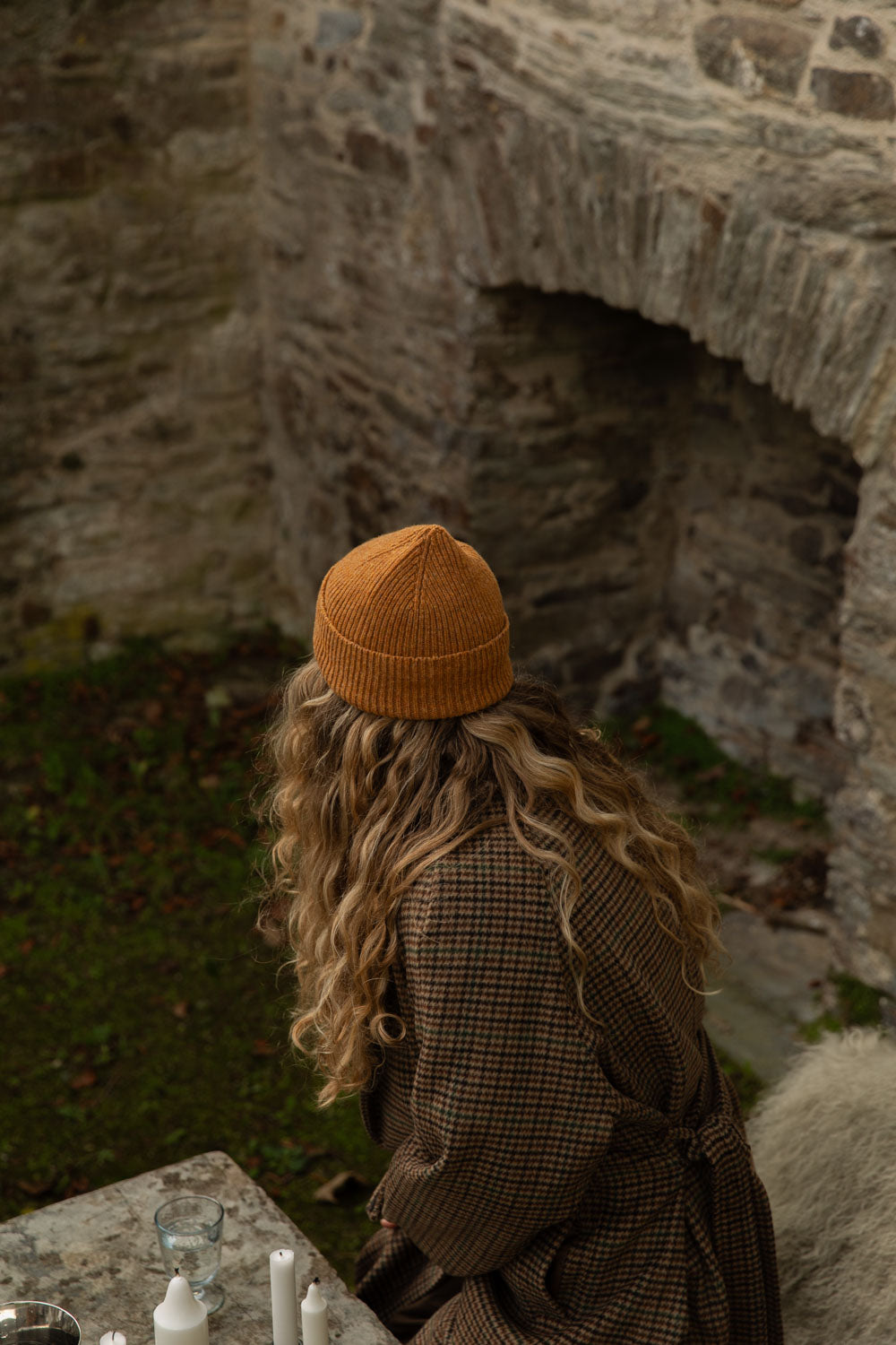 Woman in a goldcrest orange ribbed merino beanie and check coat in an old stone garden