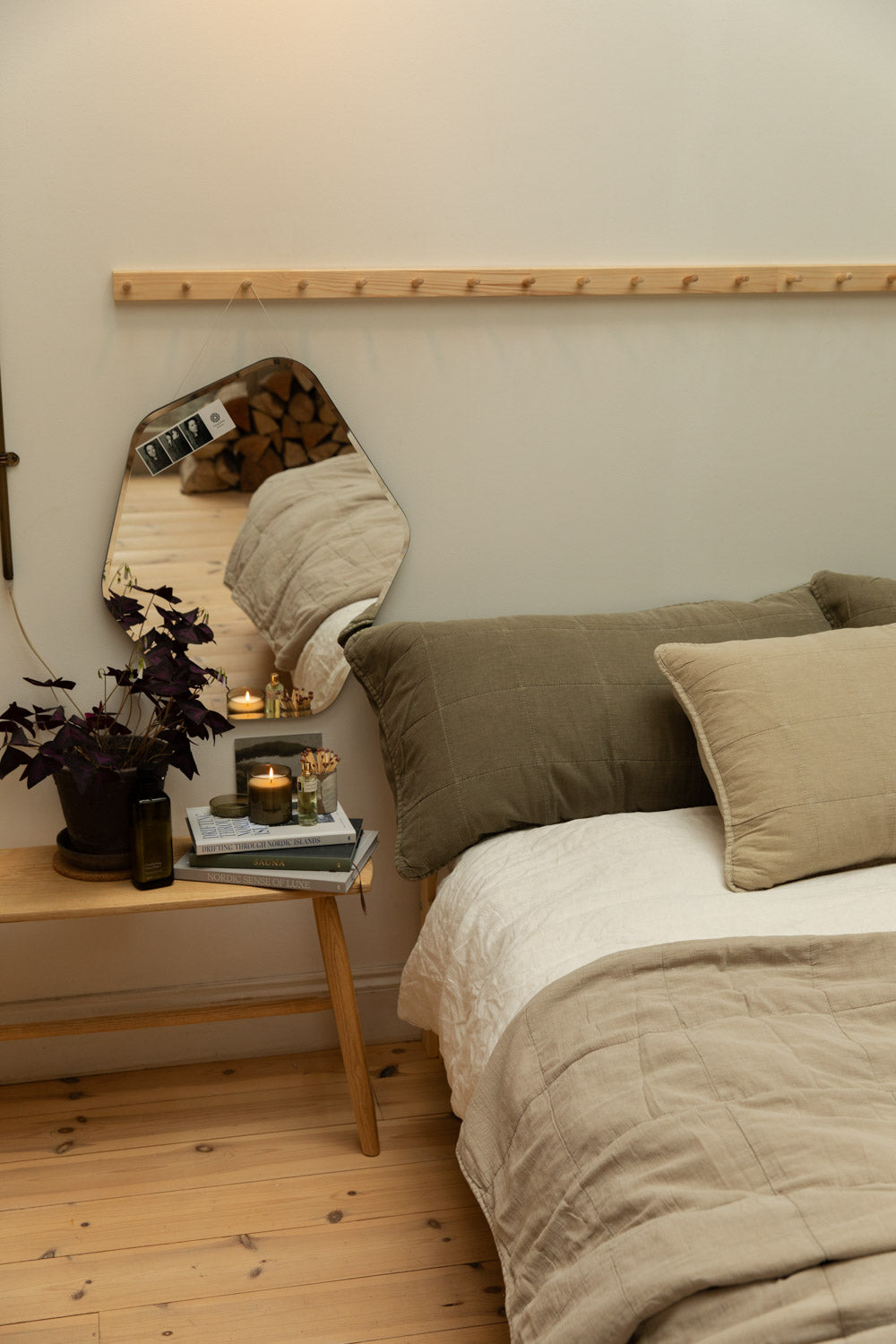 Broste Ruby Liten organic shaped mirror hanging above a pine bench bedside table styled with Nordic books, lit candle, perfume bottle and purple oxalis plant, with eucalyptus and fog quilted linen headboard cushions on a white linen bed