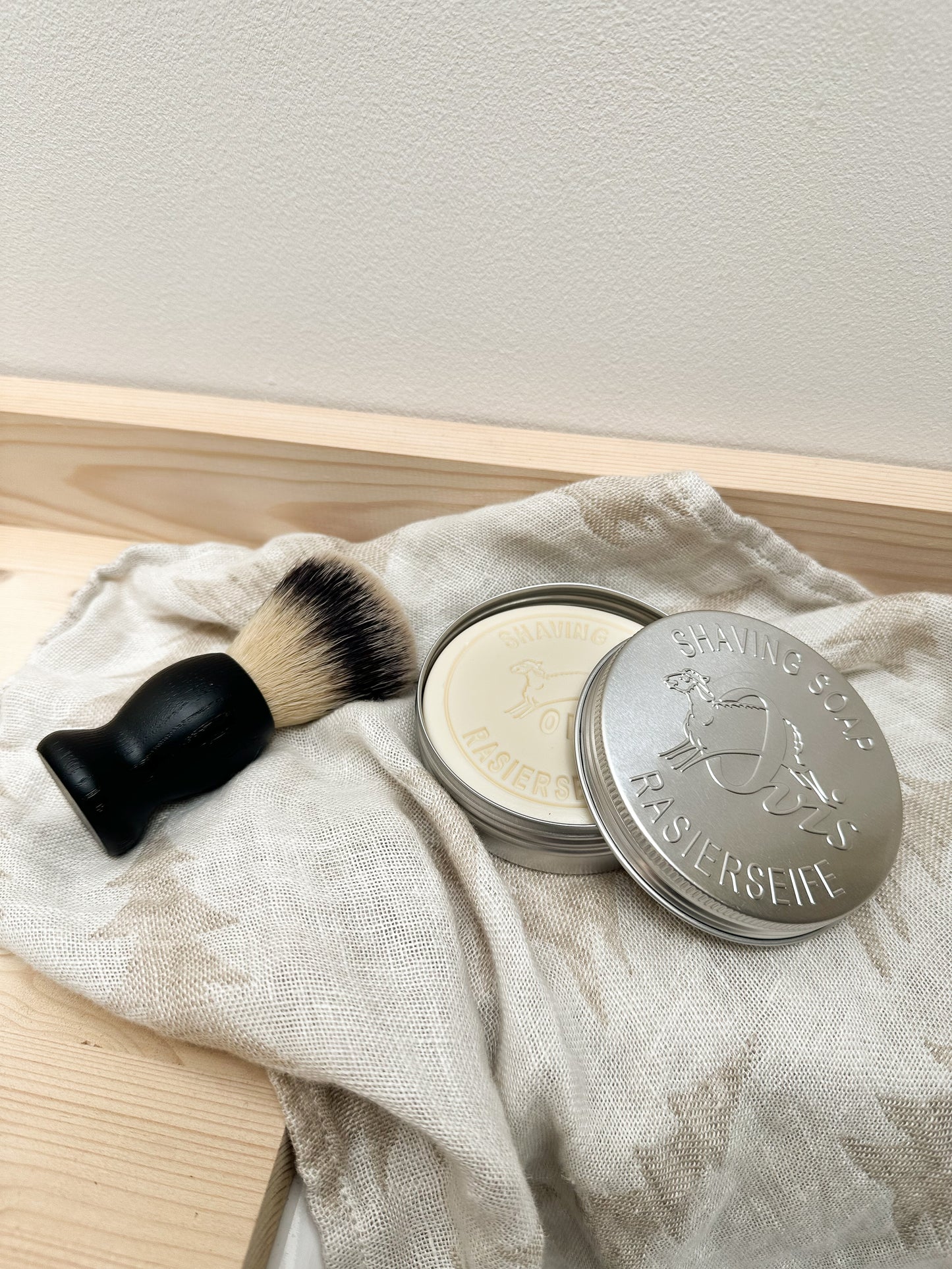 Shaving brush and silver container with shaving soap bar inside, on a light fabric with a neutral background.