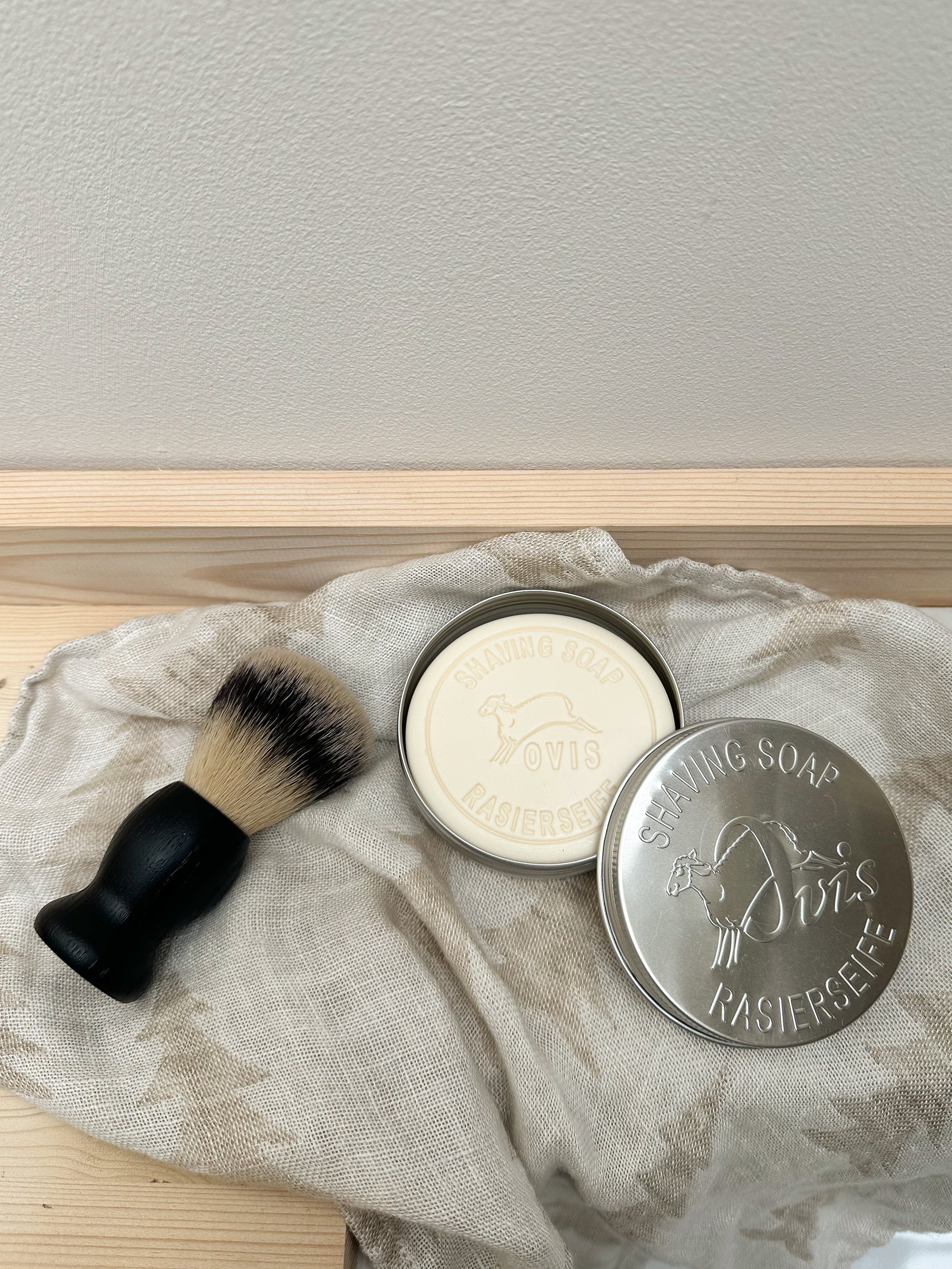 Shaving brush and shaving soap bar in an open silver tin, on a textured cloth with a neutral background