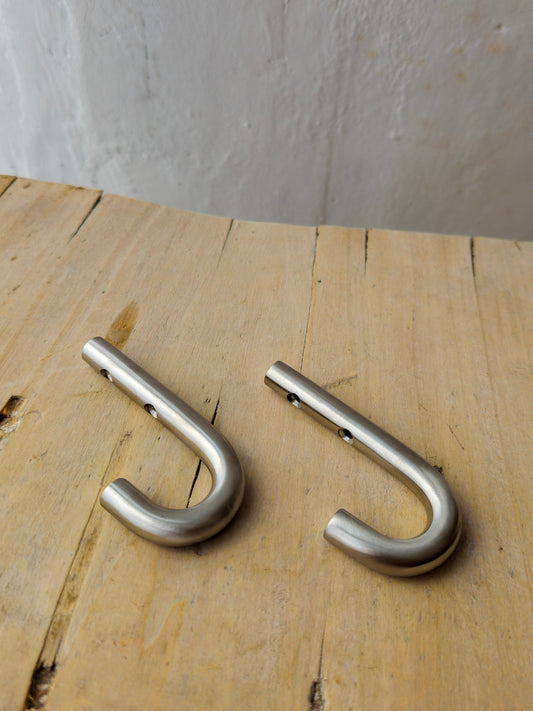 Two silver metal J-hooks with a brushed finish, displayed side-by-side on a light-colored wooden surface against a neutral gray background.