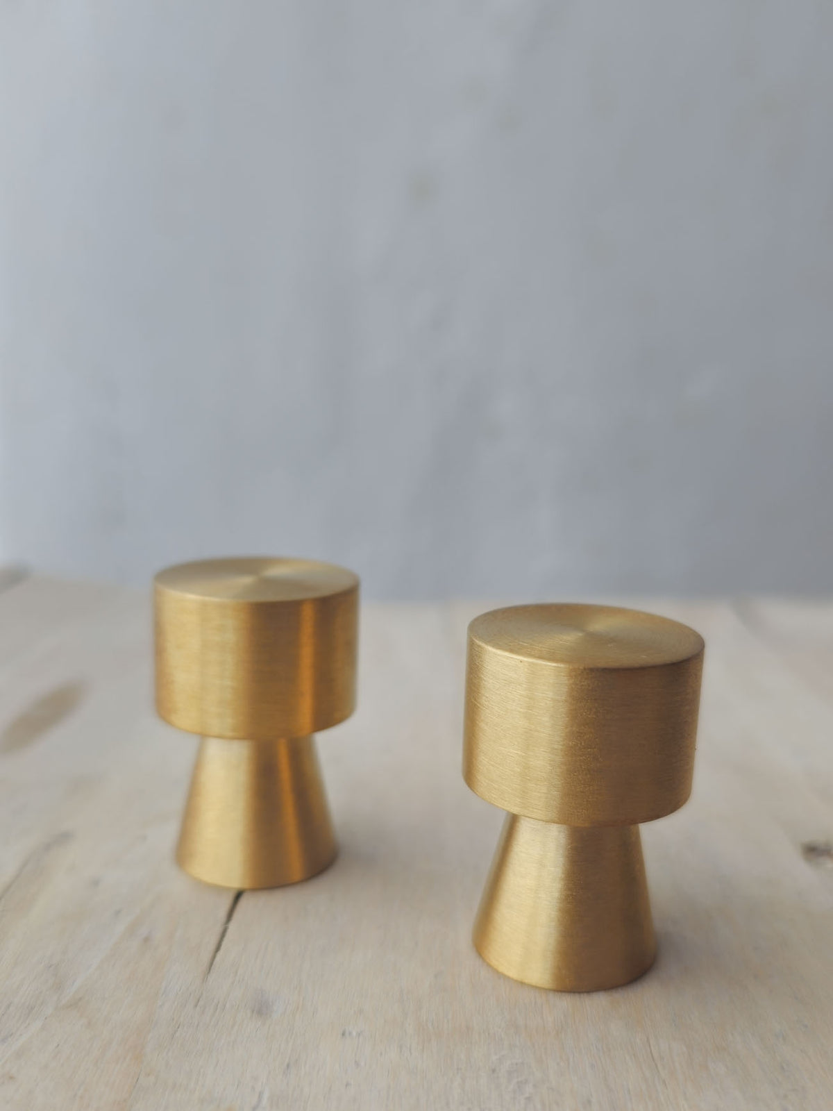 Two brushed brass cabinet knobs with a unique tapered cylindrical design, standing on a light wood surface against a soft gray background.
