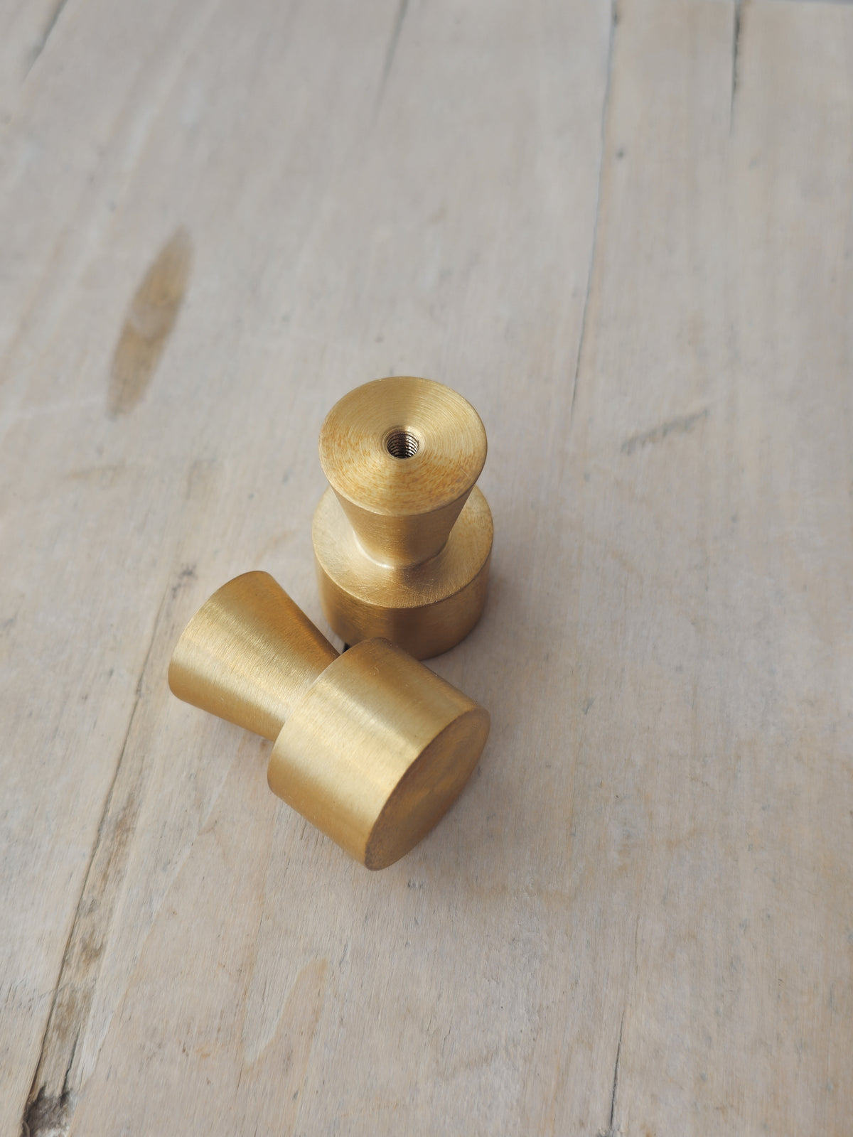 Two brass cabinet knobs with a brushed gold finish, one standing vertically and the other lying diagonally, set against a light wood grain background.