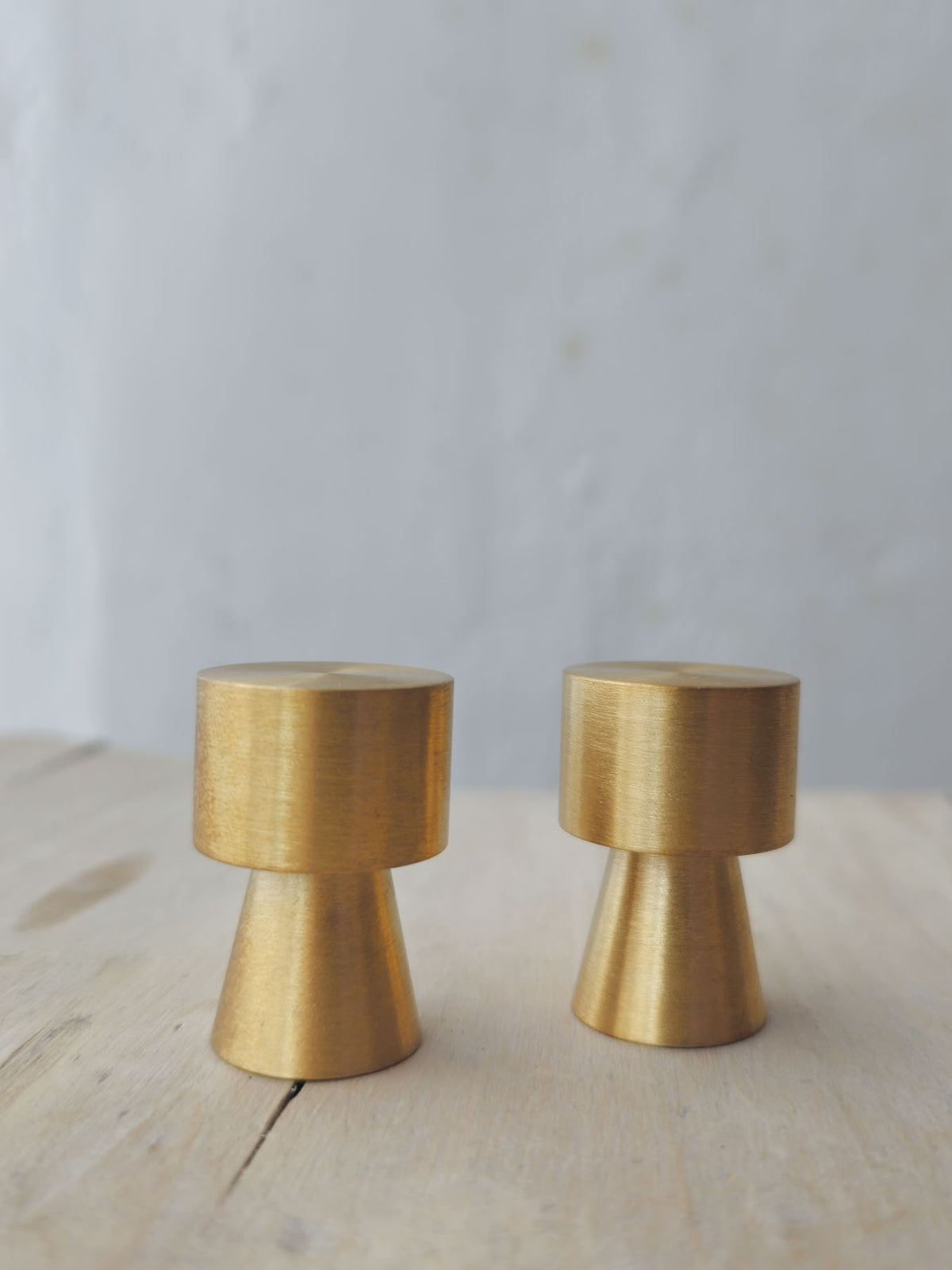 Two brushed brass cabinet knobs with a unique tapered design, displayed on a light wood surface against a soft, light gray background.