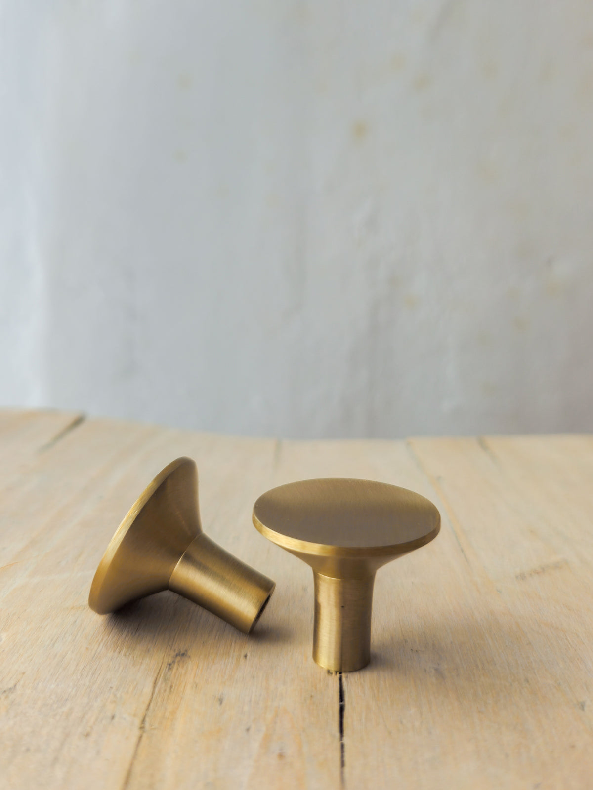 Two round cabinet knobs in a brushed brass finish, one standing upright and the other lying on its side, displayed on a light wooden surface against a neutral, softly blurred background.