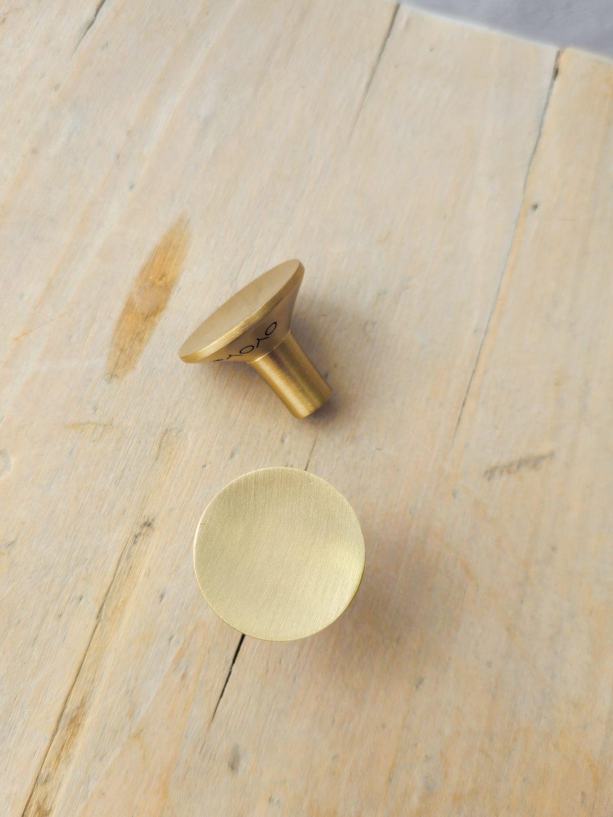 Two round cabinet knobs in a brushed brass finish, one with a flat circular face and one with a conical top, displayed on a light wood plank surface.