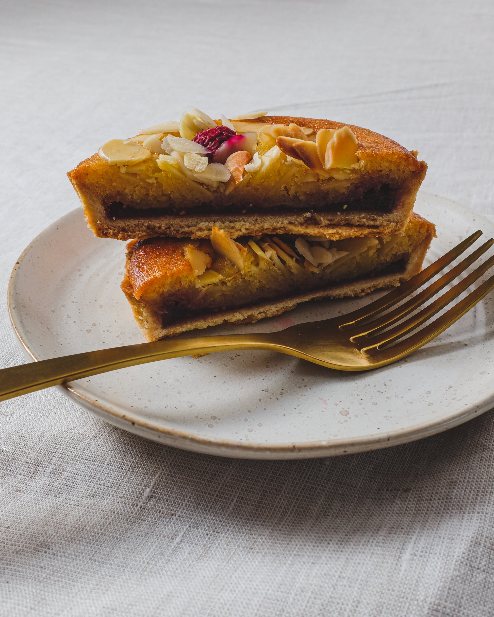 A piece of cake with nuts and a golden fork on a white cake plate with a speckled white glaze, placed on a linen tablecloth.