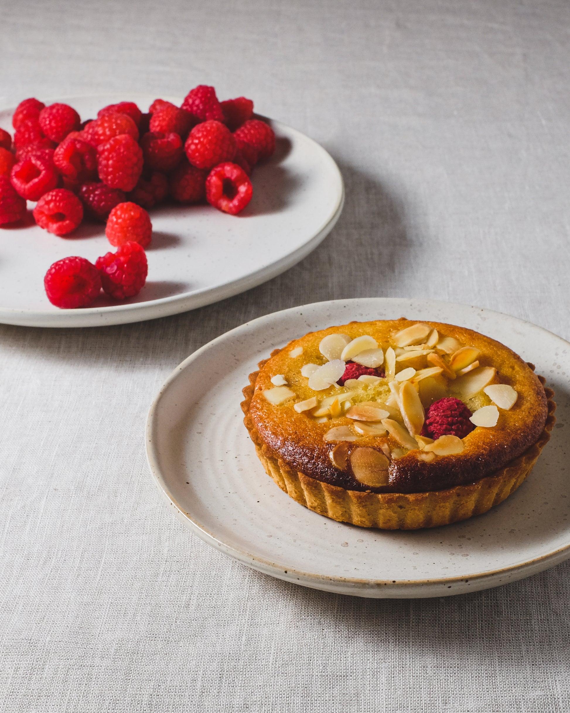 Speckled white ceramic cake plate with a tart on, with another plate in the background piled with raspberries on a light tablecloth.