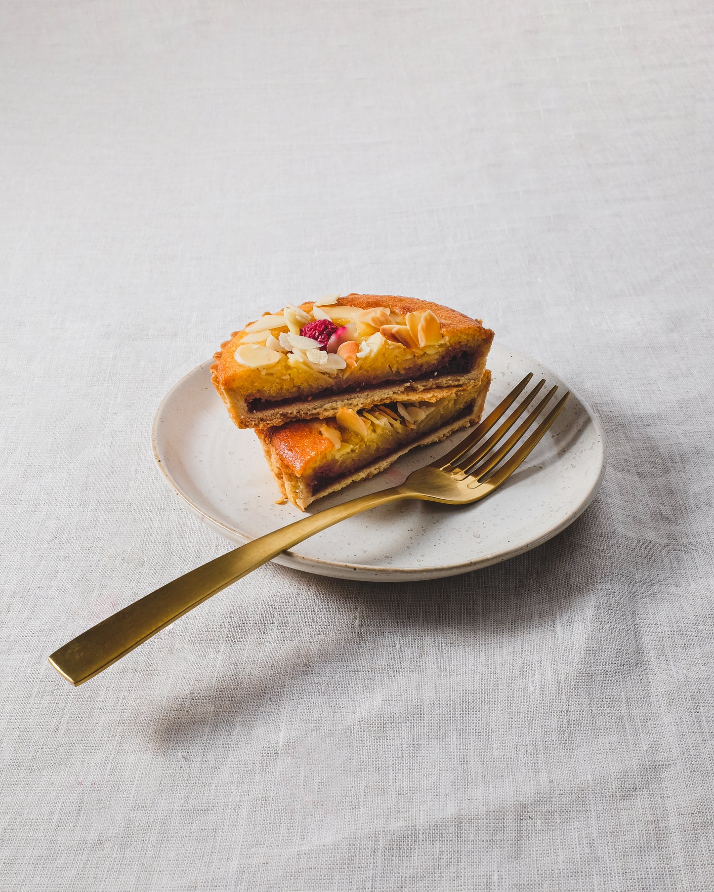 A piece of cake with nuts and a golden fork on a white cake plate with a speckled white glaze, placed on a linen tablecloth.