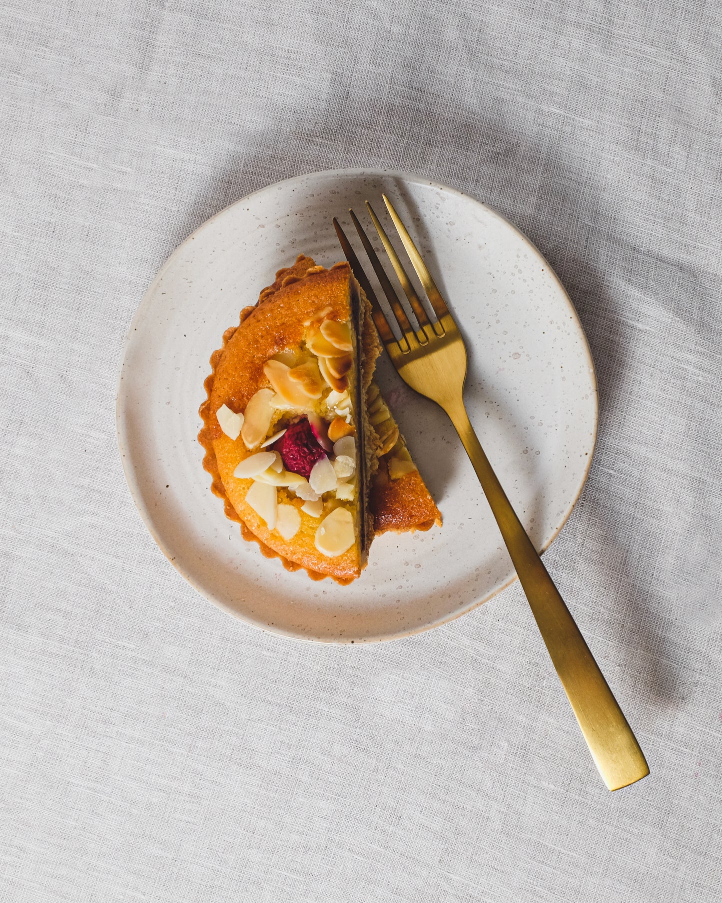 A piece of cake with nuts and a golden fork on a white cake plate with a speckled white glaze, placed on a linen tablecloth.