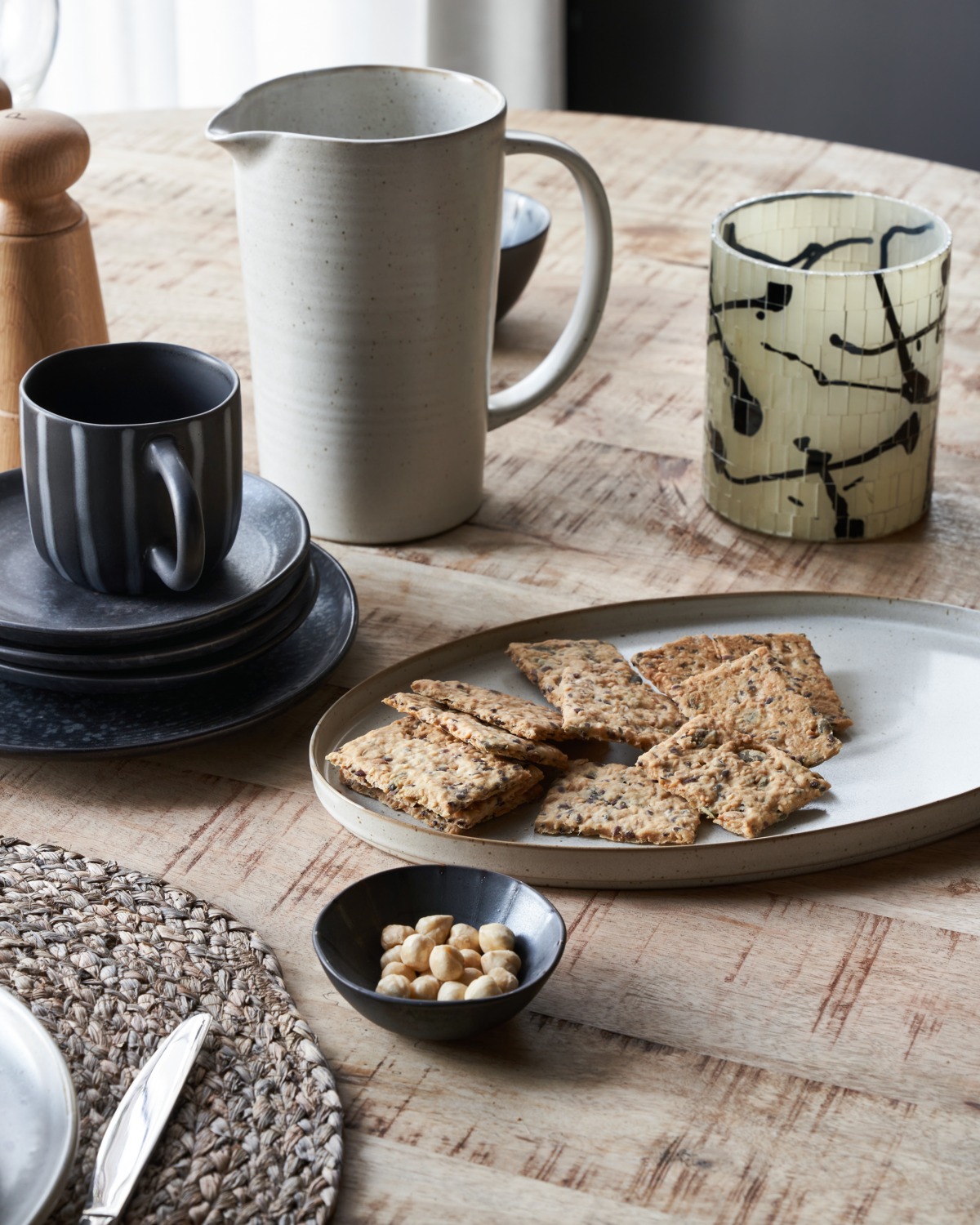 A stoneware oval serving dish on a wooden table, surrounded by various table settings including a black stoneware mug, a patterned glass, and a bowl with food items.