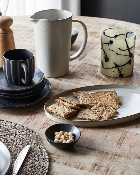 A stoneware oval serving dish on a wooden table, surrounded by various table settings including a black stoneware mug, a patterned glass, and a bowl with food items.