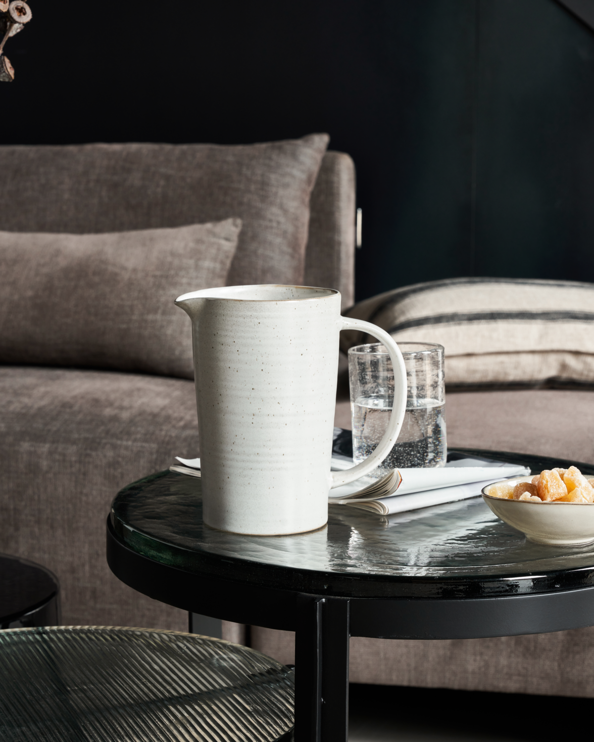White ceramic pitcher on a black round table with a dark sofa and cushions in the background.