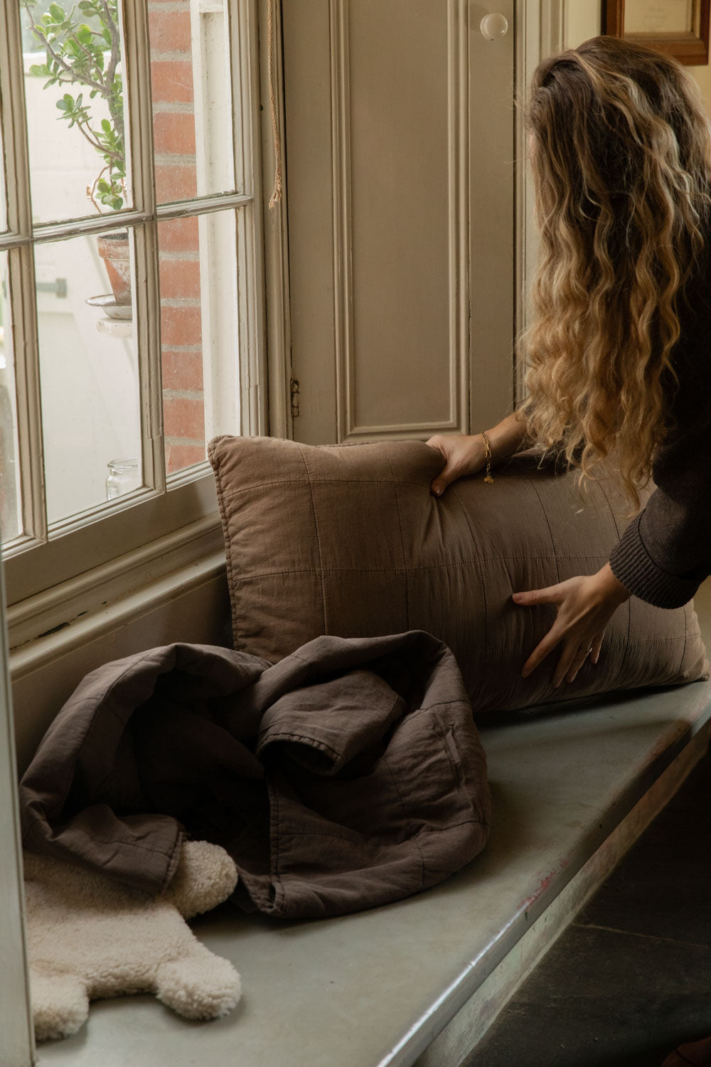 Hands setting a truffle-brown quilted cushion on a window seat beside a brown quilt and teddy hot water bottle