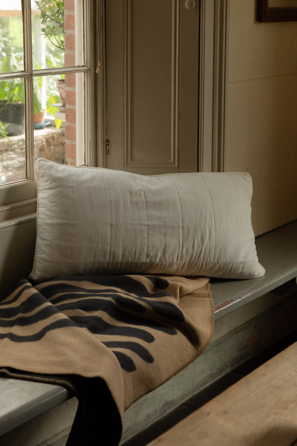 Butter Cream quilted headboard cushion on a window seat, on top of a sand and black Brita Pine lambswool blanket