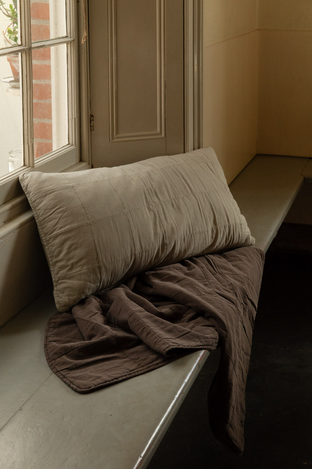 Fog quilted headboard cushion resting on a crumpled brown Soil stonewashed cotton quilt on a sunlit window seat