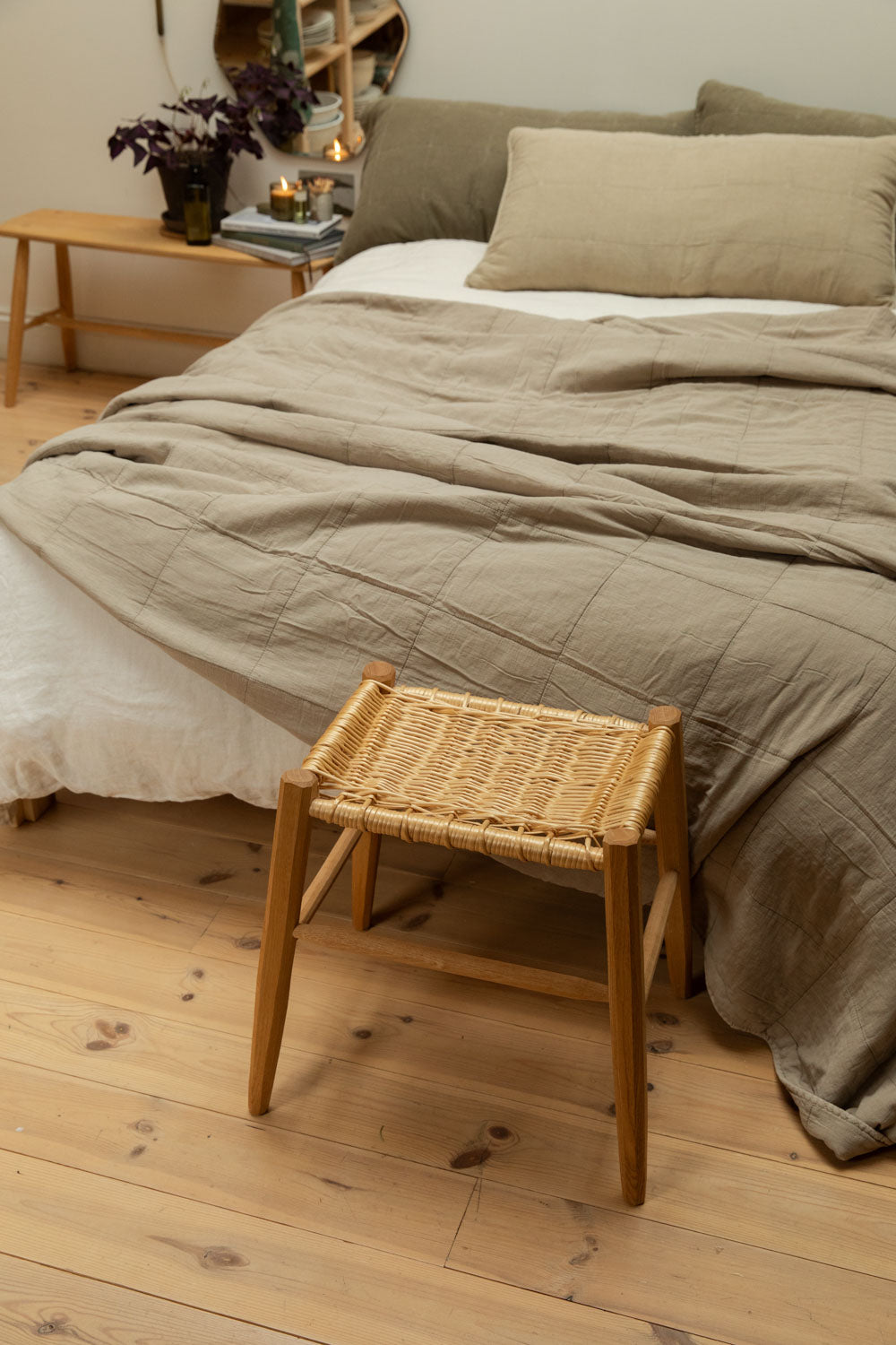 Rattan and oak stool at the foot of a bed dressed with a cotton quilt in vintage linen, pine floorboards and candlelit bedside table with dark foliage plant.