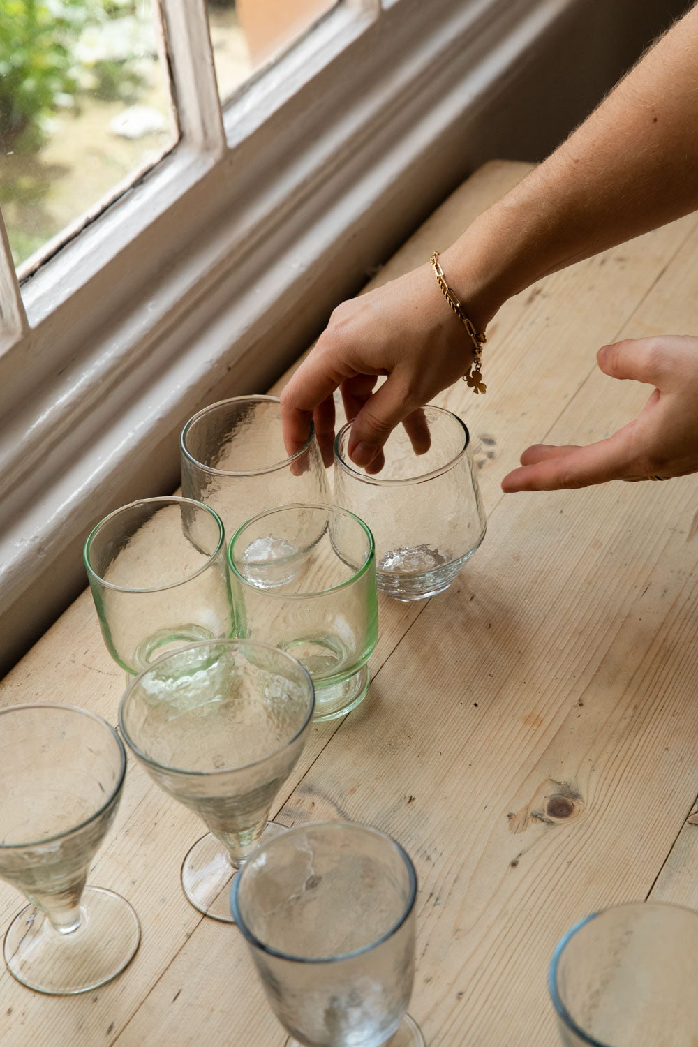 Hands arranging Meknes recycled glass tumblers and stemmed glasses on a sunny windowsill table