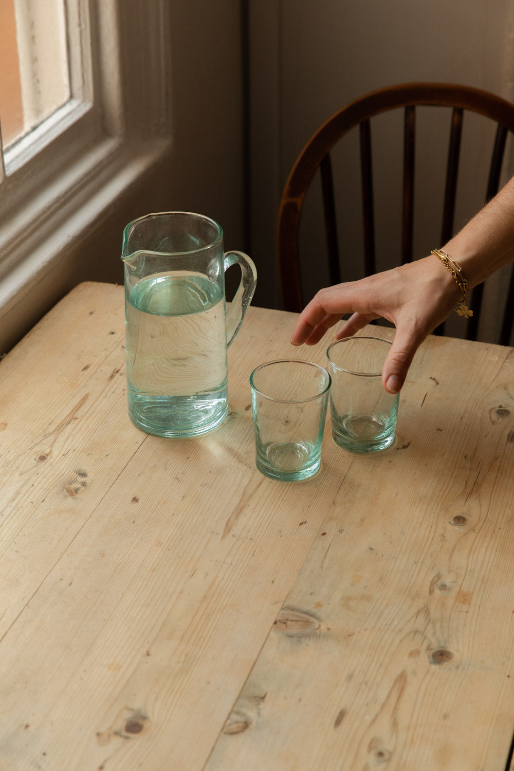 Hand-blown recycled glass jug of water beside two green-tinted tumblers on a pine table