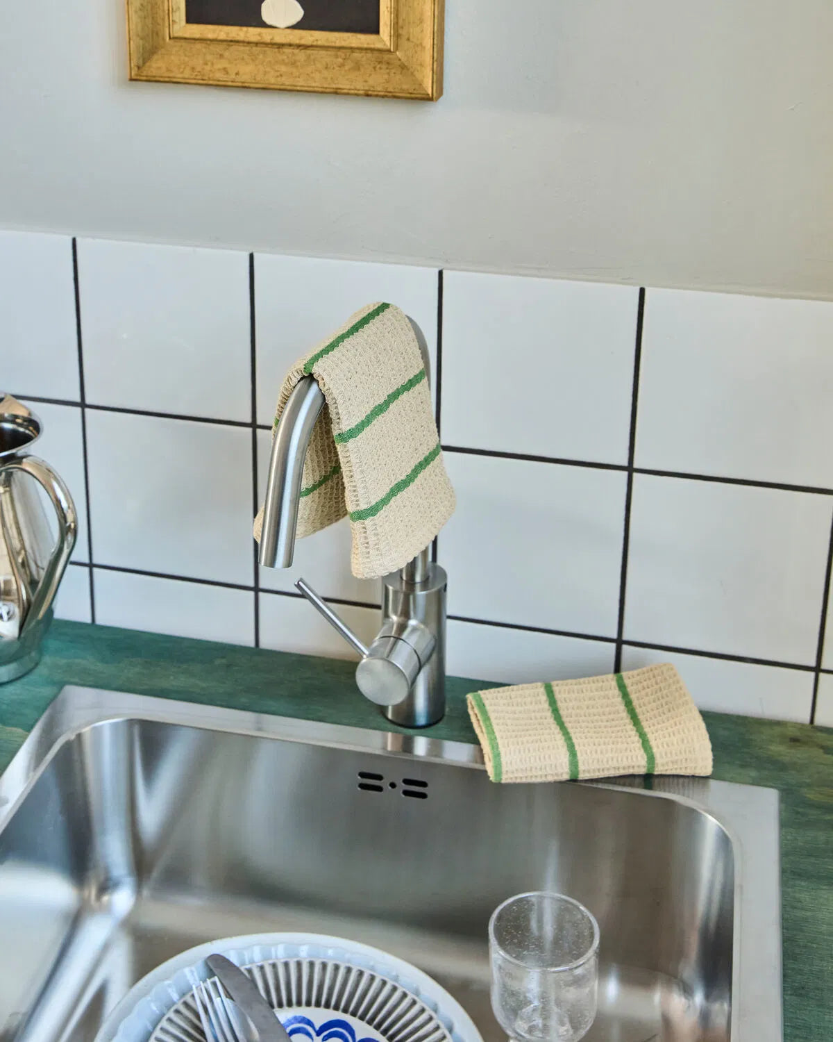 Two sand and green striped dishcloth are shown in a kitchen setting. One is draped over a silver tap, while the other is placed next to the tap on the work surface.