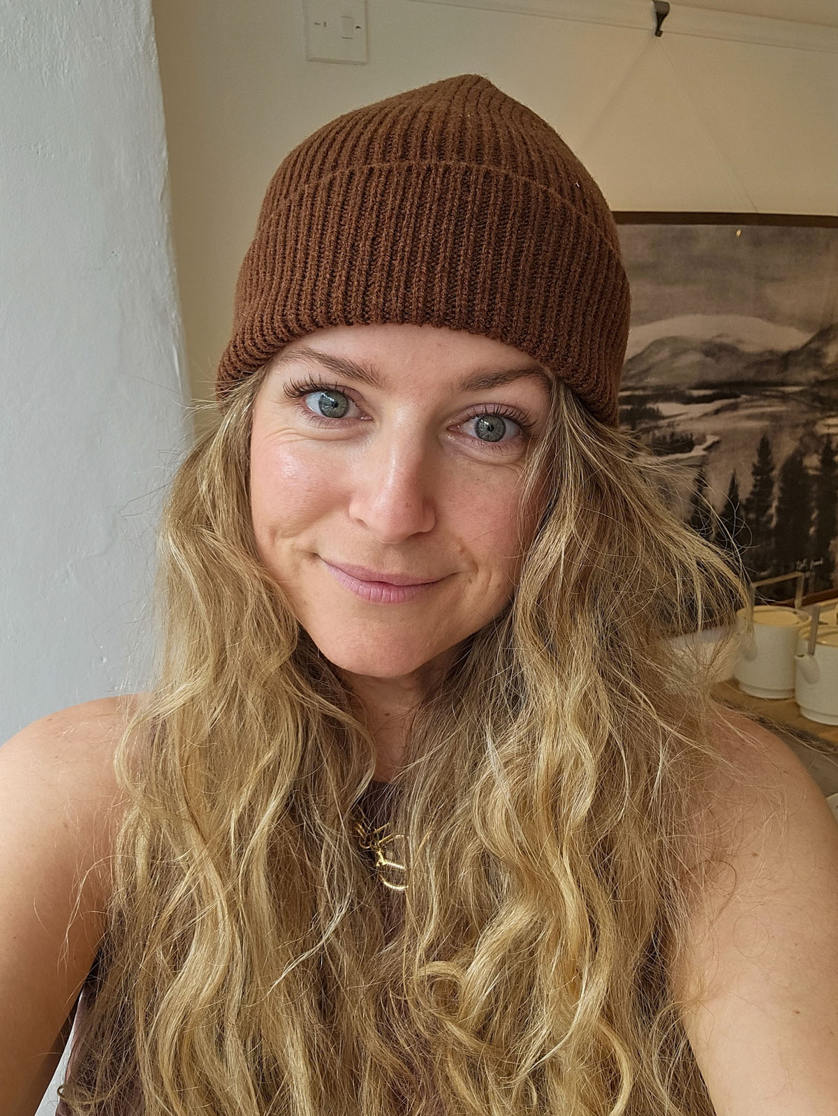 A woman with long blonde wavy hair wearing a brown ribbed knit beanie hat, photographed in an indoor setting.