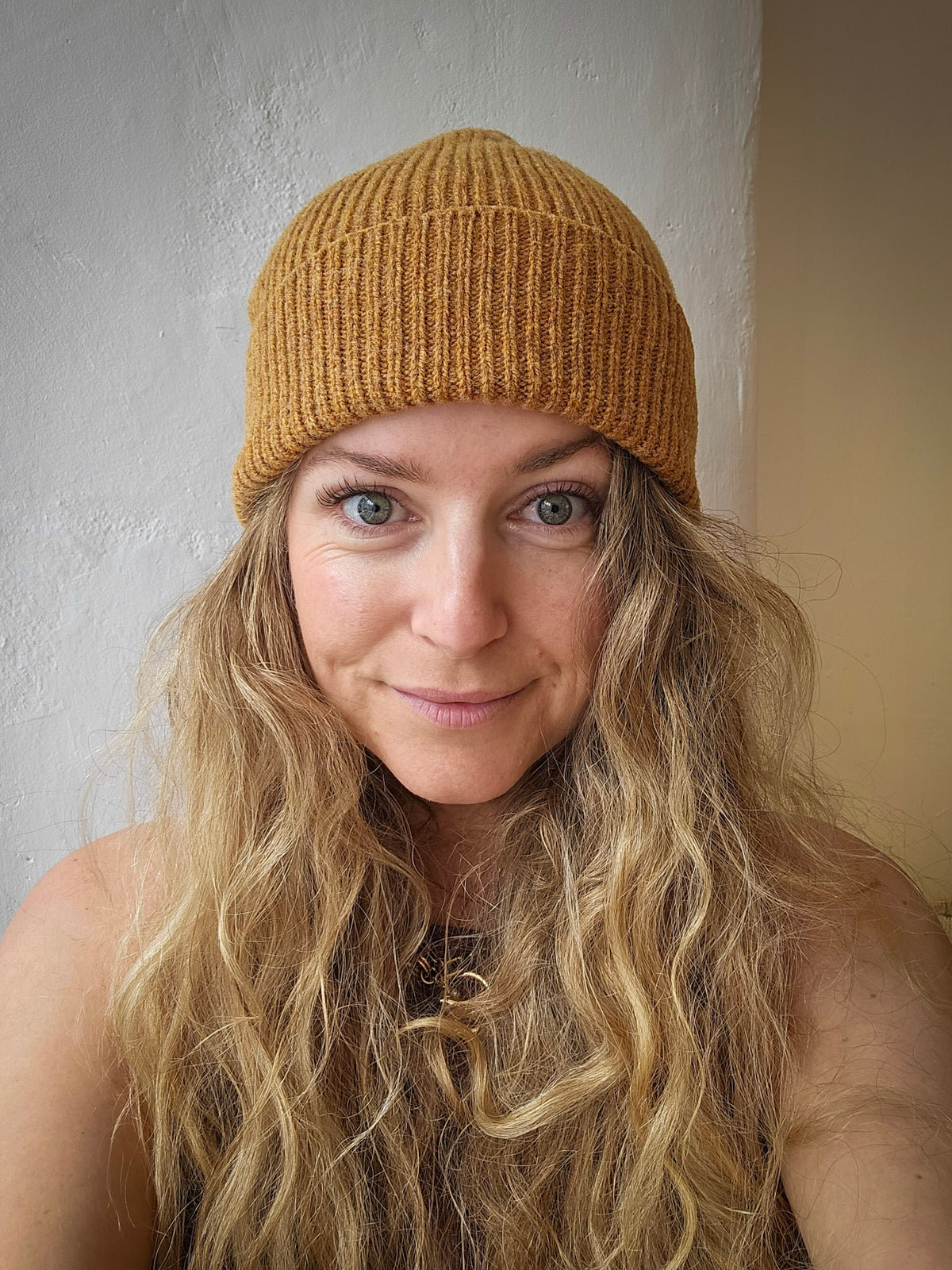 A woman with long wavy blonde hair wearing a mustard golden yellow ribbed knit beanie hat, photographed in a casual indoor setting against a textured off-white wall.