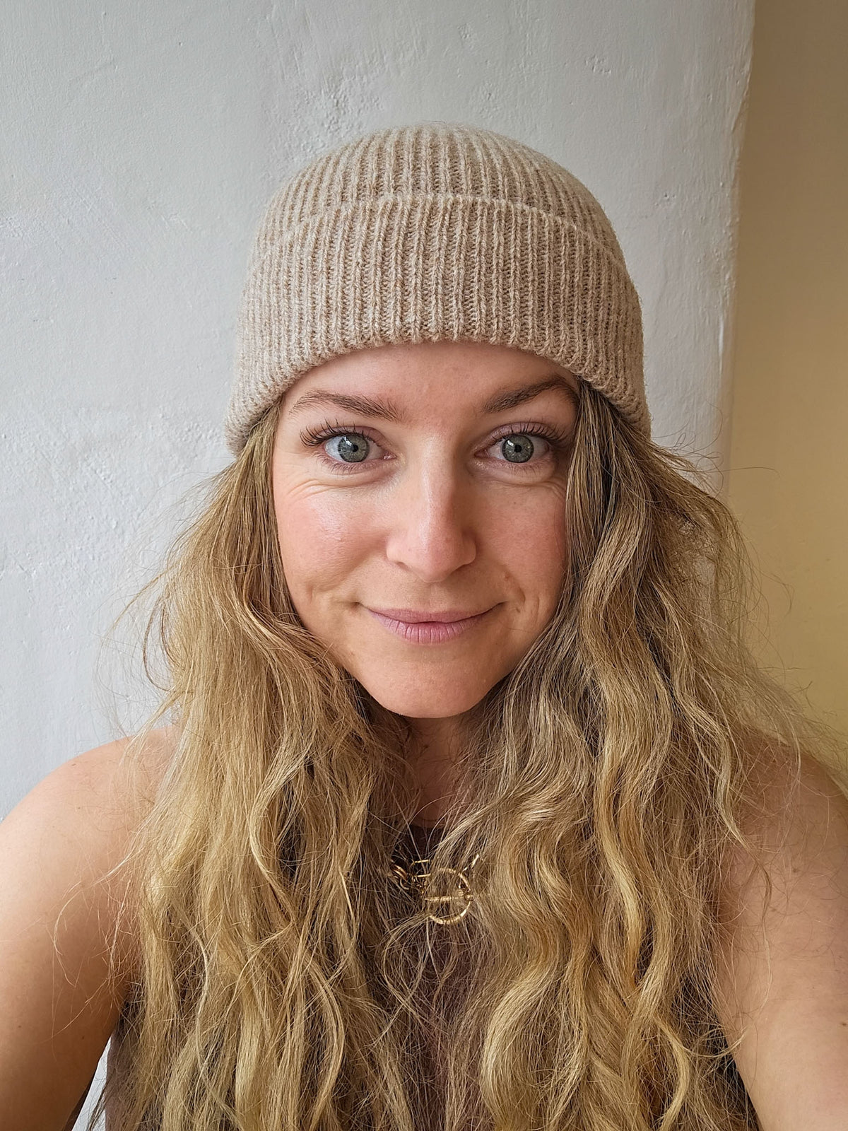 A woman with long wavy blonde hair wearing a beige ribbed knit beanie hat, photographed in a close-up portrait against a neutral indoor wall.