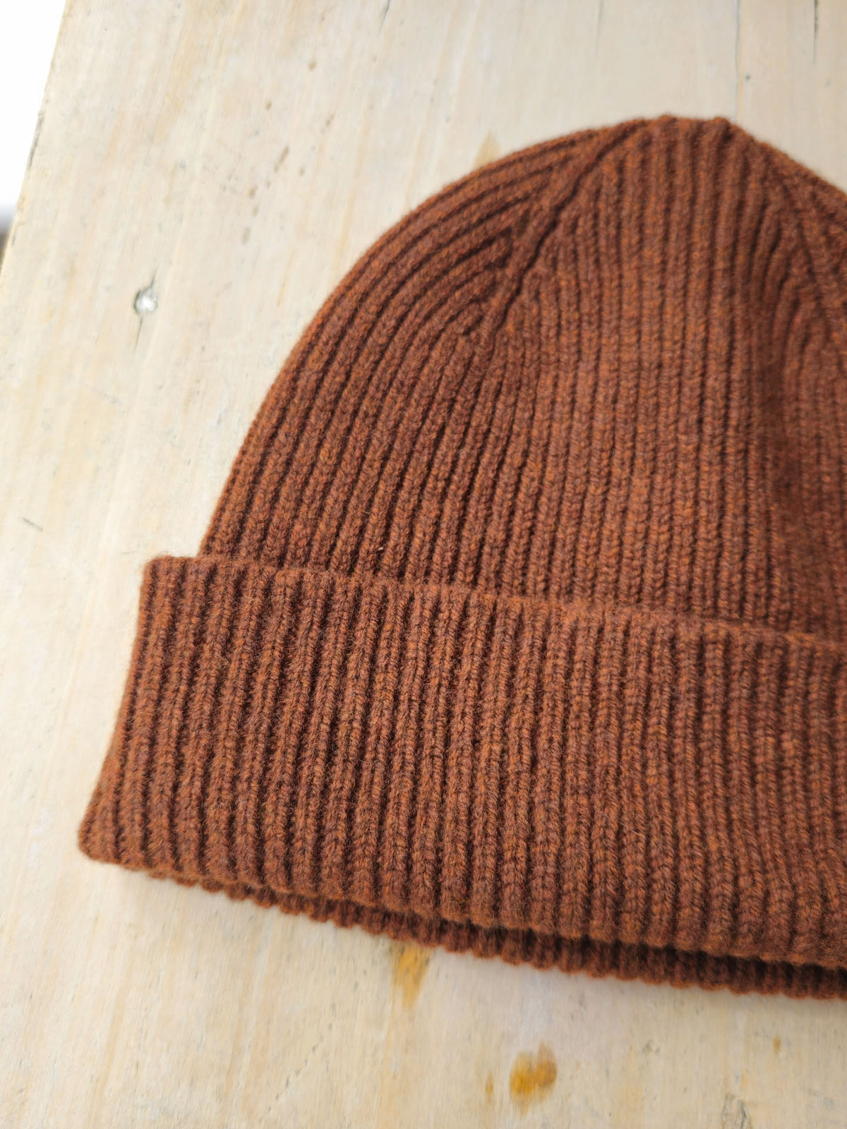 A close-up of a ribbed knit beanie in a warm rust brown color, resting on a light-colored wooden surface.