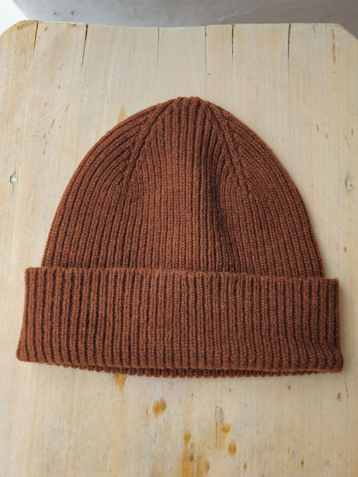 A ribbed knit beanie hat in a warm rust brown color, laid flat on a light-colored wooden surface.