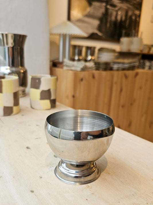 A polished silver pedestal bowl with a flared base, shown on a light wood tabletop in a softly blurred interior setting.