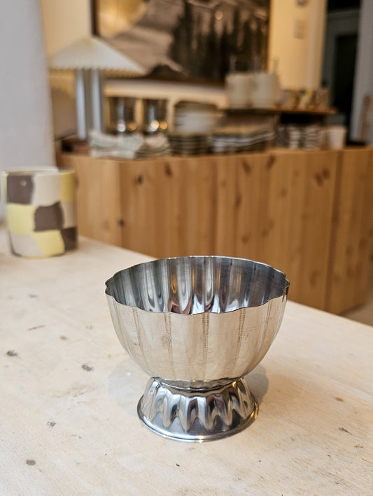 A polished silver pedestal bowl with a fluted rim, displayed on a light wooden table with a softly blurred kitchen background.