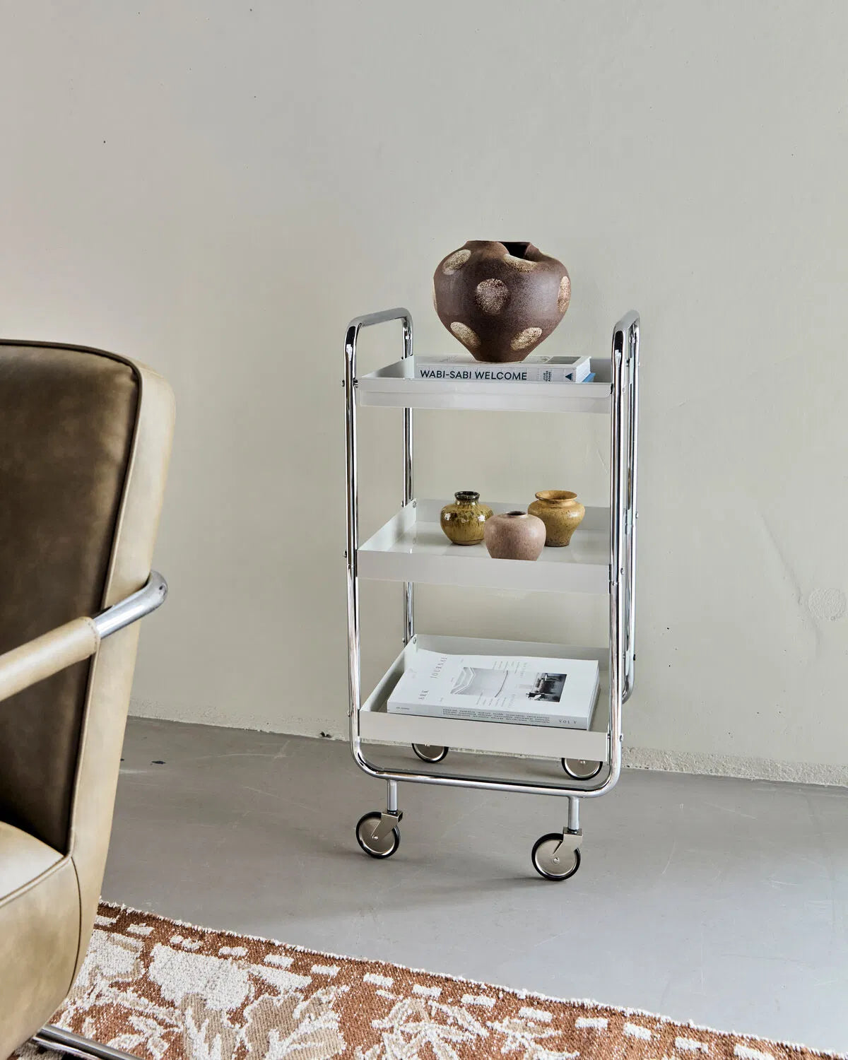 Small Roll storage trolley in white and chrome, shown on a concrete wall against an off-white painted wall. There are various objects placed onto to trolley, including vases and books.