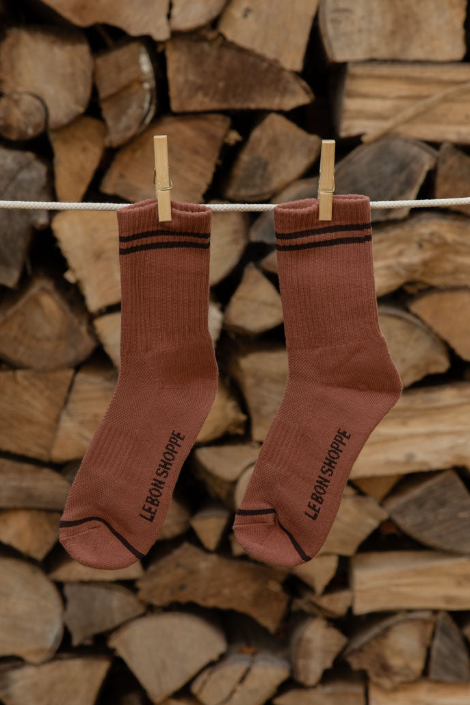 Le Bon Shoppe Boyfriend Socks in Clay, rust with dark brown stripes, pegged on a line against stacked logs