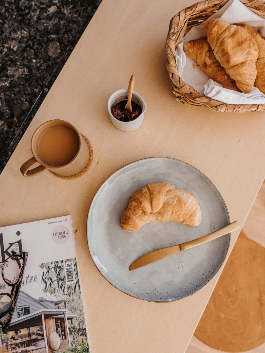 A rustic dinner plate with a bluey grey glaze, displayed on a table with a croissant, cutlery, and a magazine, indicating a stylish yet casual dining setting.