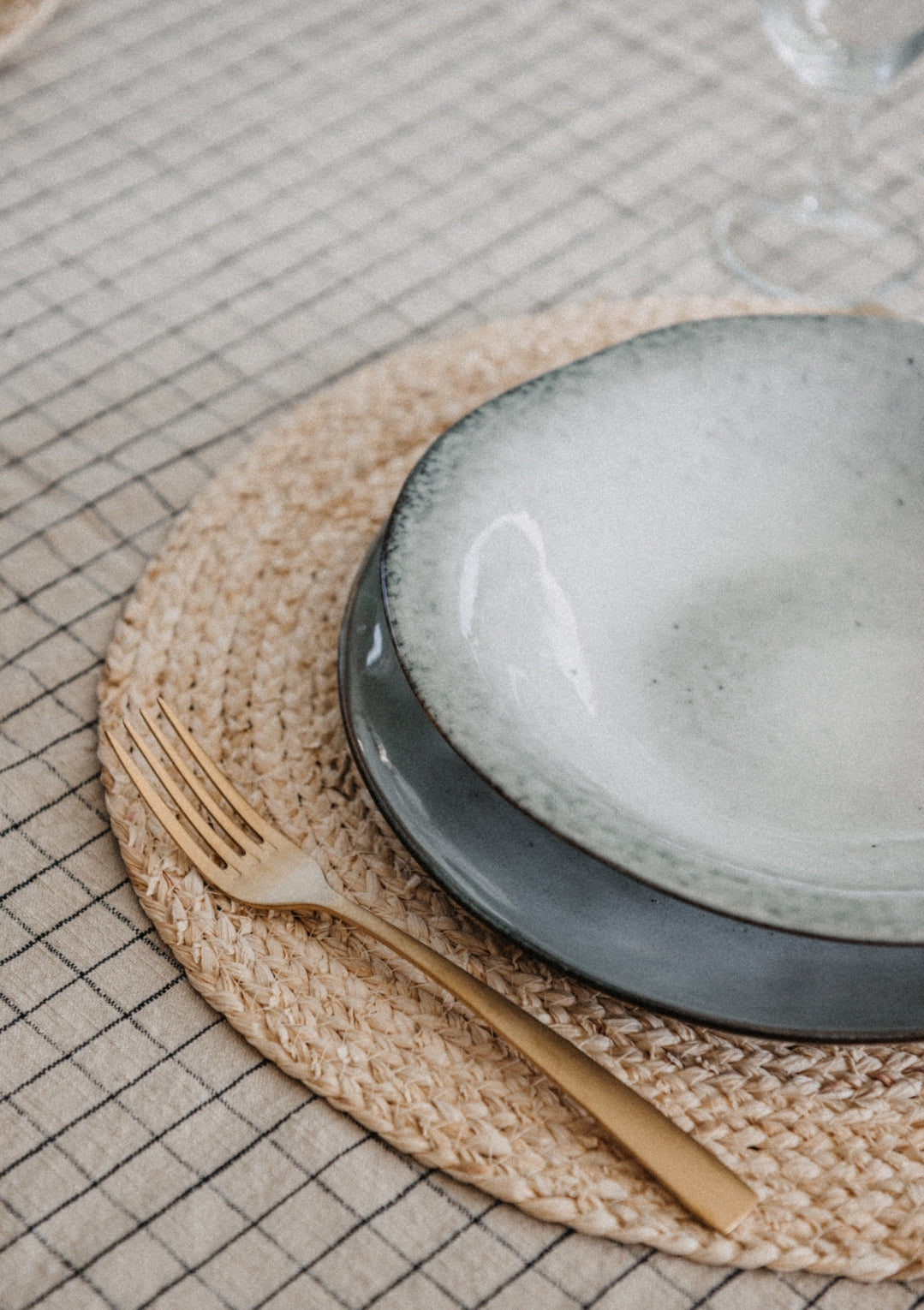 A rustic grey blue pasta bowl on a woven placemat with a fork to the left, set on a checked tablecloth.