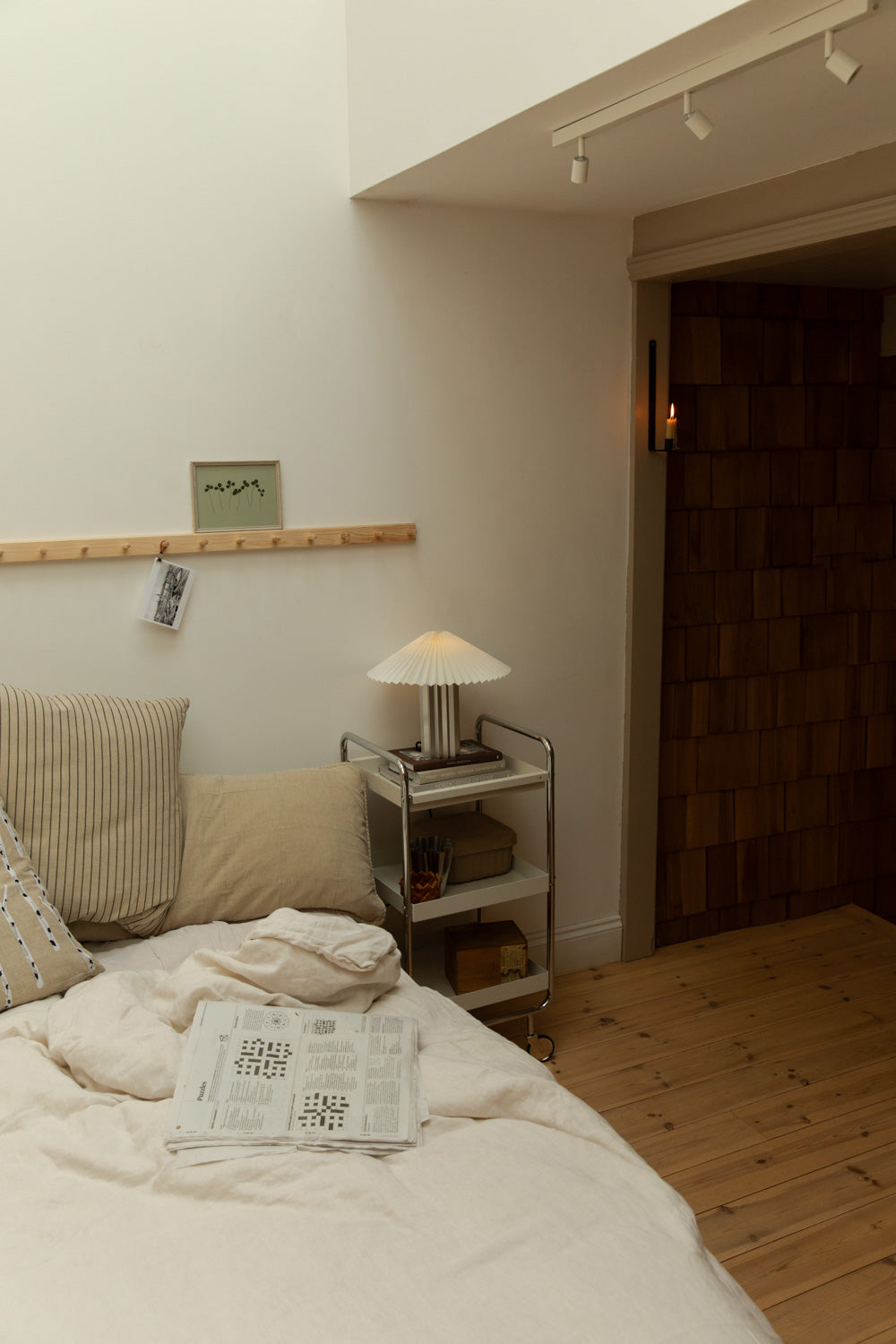 Scandi bedroom with pleated table lamp on chrome trolley, open newspaper on white linen bed, pine peg rail and botanical print.