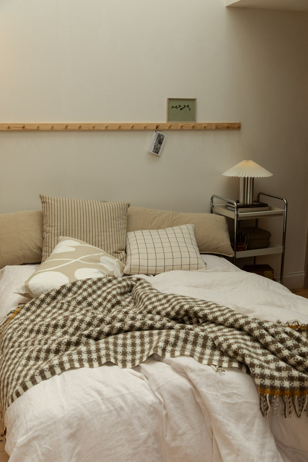 Scandi bedroom with houndstooth wool throw draped over white linen bed, pleated table lamp on chrome trolley and pine peg rail.