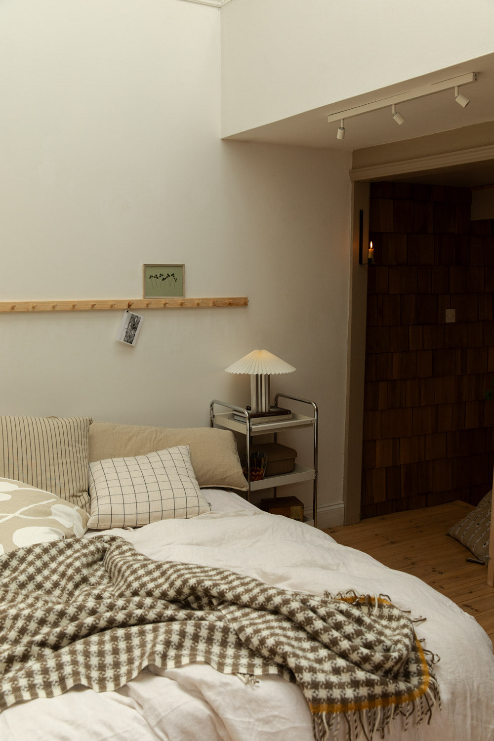 Scandi bedroom with houndstooth wool throw draped over white linen bed, pleated table lamp on chrome trolley and pine peg rail.